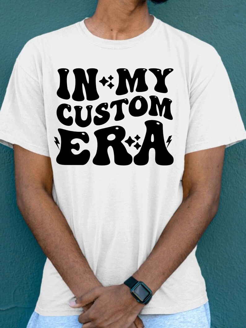 K&ouml;nnte beinhalten: Wei&szlig;es T-Shirt mit schwarzem Text, der "IN MY CUSTOM ERA" lautet, mit Sternen und Blitzen um den Text herum.