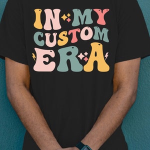 K&ouml;nnte beinhalten: Schwarzes T-Shirt mit einer bunten Grafik, auf der "In My Custom Era" mit Sternen und Blitzen steht.