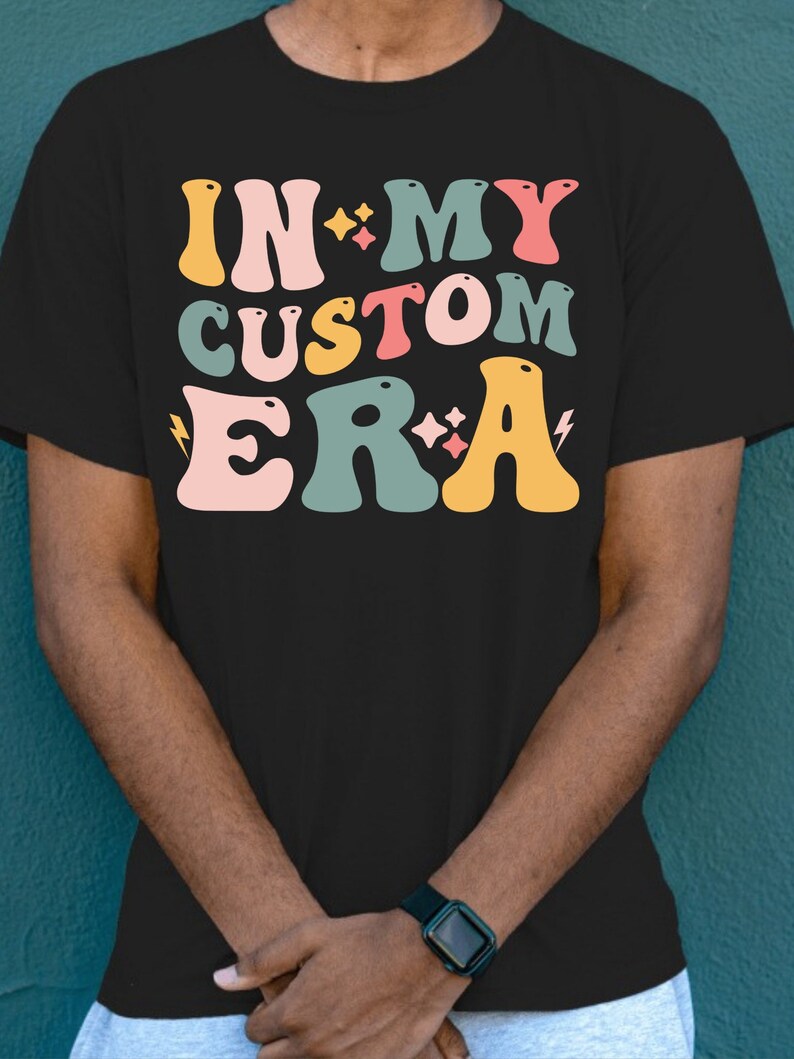 K&ouml;nnte beinhalten: Schwarzes T-Shirt mit einer bunten Grafik, auf der "In My Custom Era" mit Sternen und Blitzen steht.