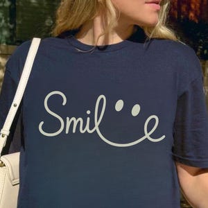 Könnte beinhalten: Ein marineblaues T-Shirt mit einem weißen, kursiv geschriebenen "Smile"-Motiv mit zwei weißen Punkten über dem "l", die ein Smiley-Gesicht bilden.