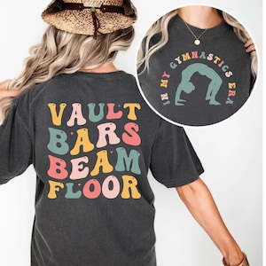 Puede incluir: Camiseta gris oscuro con texto y gráficos coloridos. La parte trasera de la camiseta presenta las palabras "VAULT BARS BEAM FLOOR" en una fuente retro. Un gráfico circular en la parte delantera dice "IN MY GYMNASTICS ERA" con una gimnasta en puente.