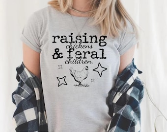 May include: A light gray t-shirt with the text "raising chickens & feral children." A black illustration of a chicken and star shapes are below the text. The shirt has rolled-up sleeves.