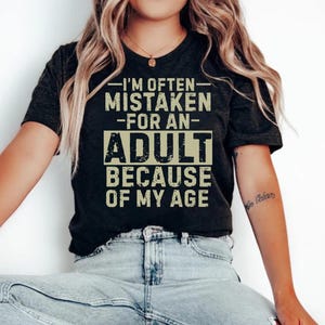 Puede incluir: Camiseta gris oscuro con el texto "I'M OFTEN MISTAKEN FOR AN ADULT BECAUSE OF MY AGE" en una fuente desgastada. La camiseta la lleva una persona con vaqueros azules.