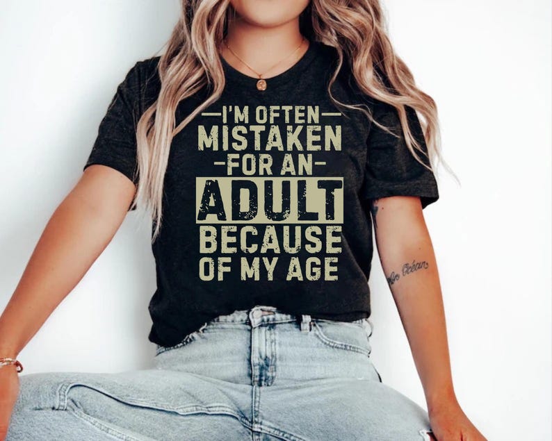 Puede incluir: Camiseta gris oscuro con el texto "I'M OFTEN MISTAKEN FOR AN ADULT BECAUSE OF MY AGE" en una fuente desgastada. La camiseta la lleva una persona con vaqueros azules.