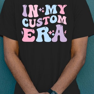 K&ouml;nnte beinhalten: Schwarzes T-Shirt mit dem Text "IN MY CUSTOM ERA" in rosa, blauen und violetten Buchstaben mit Glitzerakzenten.