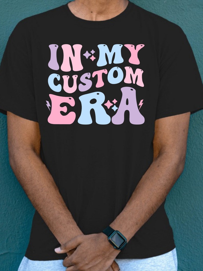 K&ouml;nnte beinhalten: Schwarzes T-Shirt mit dem Text "IN MY CUSTOM ERA" in rosa, blauen und violetten Buchstaben mit Glitzerakzenten.