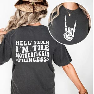 May include: Dark gray t-shirt with the text "HELL YEAH I'M THE MOTHERF*CKIN PRINCESS" in white. The back of the shirt features the same text. A close-up shows a white skeleton hand gesture. The shirt is a casual, unisex garment.