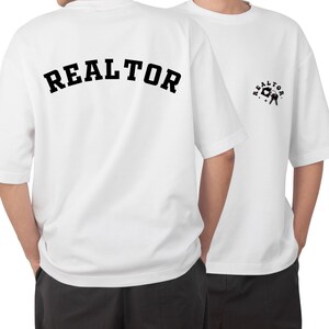 以下が含まれることがあります： 白地に黒で「REALTOR」とプリントされたTシャツ。前面には、鍵を持った家の小さな黒いグラフィックが描かれている。