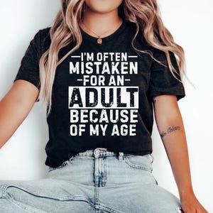 Puede incluir: Camiseta gris oscuro con el texto blanco: "I'M OFTEN MISTAKEN FOR AN ADULT BECAUSE OF MY AGE". La camiseta tiene cuello redondo y mangas cortas.