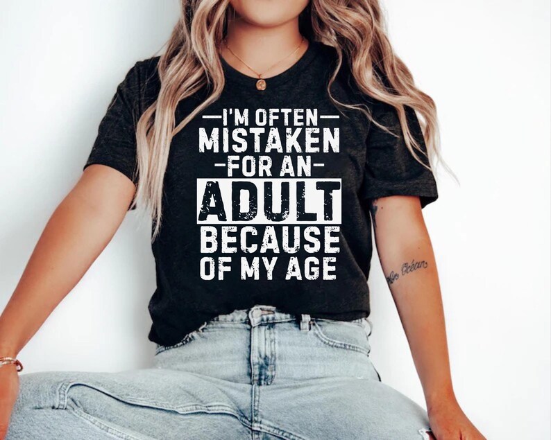 Puede incluir: Camiseta gris oscuro con el texto blanco: "I'M OFTEN MISTAKEN FOR AN ADULT BECAUSE OF MY AGE". La camiseta tiene cuello redondo y mangas cortas.