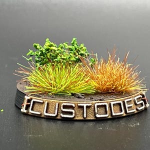 May include: A miniature model base with green and brown tufts of grass and the word "CUSTODES" written on the front.