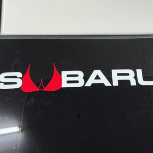 Puede incluir: Superficie negra con la palabra "SUBARU" en blanco y un gráfico rojo. El gráfico es un diseño estilizado. La superficie tiene un borde blanco en la parte superior y un borde negro a la izquierda.