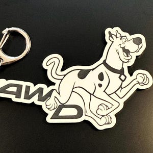 May include: A keychain featuring a cartoon dog with black spots, a black collar, and the text "AWD" in black. The dog is running and smiling. The keychain has a silver clasp.