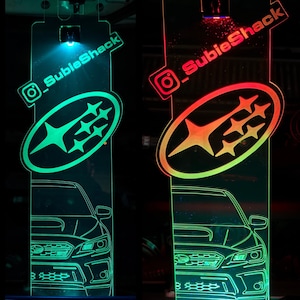 May include: Two illuminated acrylic Subaru car signs. One glows green, the other red and orange. Each features the Subaru logo and a car outline. The text "@_SubieShack" is displayed on each sign.