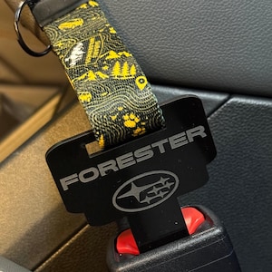 Pode incluir: Fivela de cinto de segurança preta com a palavra "FORESTER" e um logotipo da Subaru. A fivela está presa a uma alça estampada com desenhos de montanhas e florestas amarelas e pretas. O cinto de segurança está afivelado.