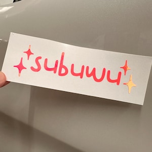 Puede incluir: Una pegatina blanca con la palabra "subuwu" escrita en rosa con un efecto brillante. Hay cuatro estrellas rosas en la pegatina, dos a cada lado de la palabra.