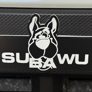 May include: A black and white sticker featuring a cartoon dog's face above the word "SUBAWU" in white capital letters. The sticker is on a textured black surface.
