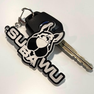 May include: A black and white Subaru keyring with the word "Subaru" in bold, white letters. The keyring features a cartoon graphic of a bulldog. A car key is next to the keyring.