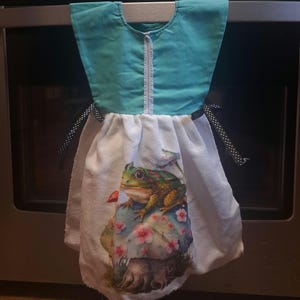 May include: A teal and white kitchen towel with a frog and mushroom design. The top portion is teal with a white zipper-like trim. The bottom is white with a colorful frog illustration on a mushroom with pink flowers. Black and white polka dot ribbons are attached.