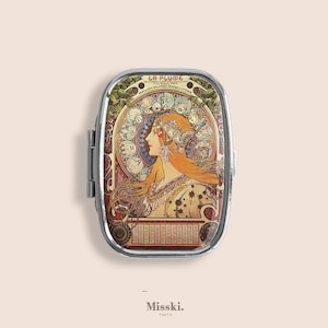 Op de afbeelding: Een zilveren pillendoosje met een Art Nouveau-stijl illustratie van een vrouw met golvend rood haar en een bloemenhoofddeksel. De doos heeft de tekst "La Plume" bovenaan en de merknaam "Misski. Tokyo" onderaan.