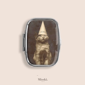 May include: A silver-framed pill box featuring a sepia-toned image of a cat wearing a dunce cap. The word "DUNCE" is printed on the cap. The cat is centered in the frame, with the Misski. Tokyo logo at the bottom.