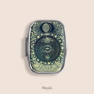 Puede incluir: Un pastillero plateado con un diseño celestial. La tapa presenta una ilustración detallada de una luna, estrellas y un ojo dentro de un patrón circular. El texto "Misski. Tokyo" está en la parte inferior.