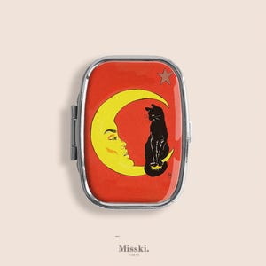 Op de afbeelding: Een rechthoekige, zilveromrande pillendoos met een rode achtergrond. De doos heeft een gele maan met een gezicht, een zwarte kat en een ster. De tekst "Misski. Tokyo" staat onderaan.