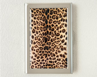 May include: A framed print featuring a leopard print pattern. The print has a silver-colored frame. The leopard print has a tan background with dark brown spots. The frame is rectangular and is displayed against a white wall.