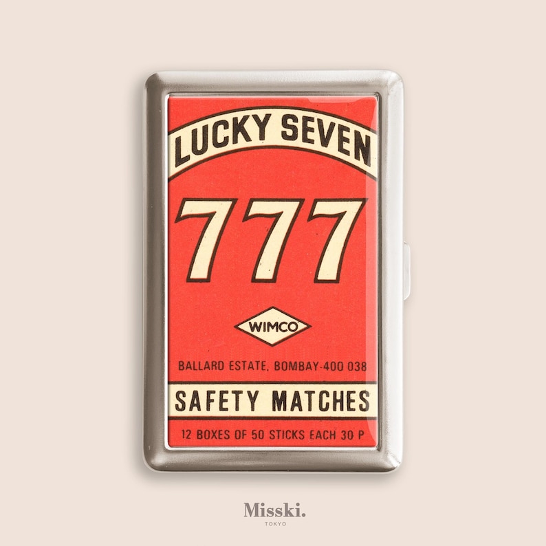 May include: A rectangular silver match case with a red and white graphic. The graphic reads "LUCKY SEVEN" above the number "777". Below the number is the text "SAFETY MATCHES". The case is from "WIMCO".