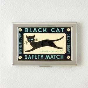 Könnte beinhalten: Ein rechteckiges Etui aus silberfarbenem Metall mit einer Vintage-Grafik einer schwarzen Katze mit türkisfarbenen Augen. Der Text lautet BLACK CAT, SAFETY MATCH, MADE IN POLAND, IMPREGNATED und AVERAGE CONTENTS 50 auf cremefarbenem und schwarzem Grund.