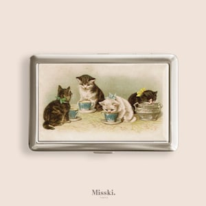 May include: A silver metal business card holder with a vintage illustration of four kittens gathered around teacups. The kittens are various colors, including black, brown, and white, and are adorned with bows. The text "Misski. Tokyo" is at the bottom.
