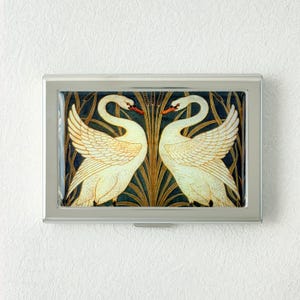 May include: A silver business card holder featuring a decorative design of two white swans facing each other. The swans have orange beaks and are set against a dark blue and gold background.
