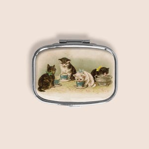 May include: An oval metal pill box featuring a printed illustration of four kittens interacting with small blue teacups and a bowl. The kittens are depicted in various colours including brown tabby, white, and black, with some wearing small bows.