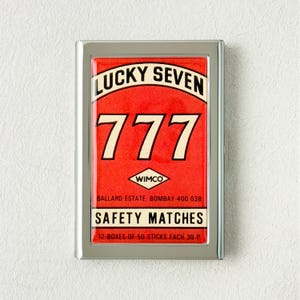 May include: A silver-framed matchbox with a red face. The text "LUCKY SEVEN" arches across the top, with the number "777" in large white digits below. The words "SAFETY MATCHES" are also visible. The matchbox contains 12 boxes of 50 matches.