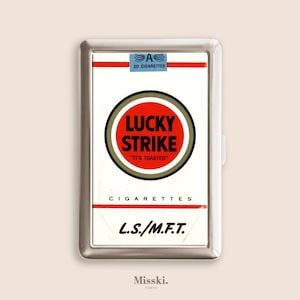 May include: A silver cigarette case featuring a Lucky Strike design. The case has a white background with the Lucky Strike logo in a red circle, and the text "IT'S TOASTED". The case also has the text "CIGARETTES" and "L.S./M.F.T.".