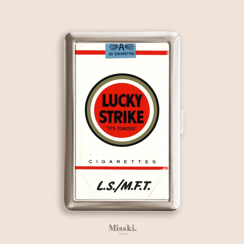 May include: A silver cigarette case featuring a Lucky Strike design. The case has a white background with the Lucky Strike logo in a red circle, and the text "IT'S TOASTED". The case also has the text "CIGARETTES" and "L.S./M.F.T.".