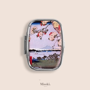 May include: A silver pill case with a hinged lid, featuring a Japanese landscape print. The artwork depicts a mountain, lake, boats, and flowering branches. The case has the text "Misski. Tokyo" at the bottom.