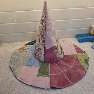 Op de afbeelding: Een handgemaakte heksenhoed met een patchwork-ontwerp. De conische kroon is gemaakt van bloemenstof en effen roze materiaal. De rand is een patchwork van verschillende stoffen in roze, blauwe, groene en beige tinten. De hoed is waarschijnlijk voor kostuum- of decoratieve doeleinden.