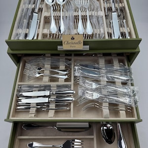 May include: A multi-tiered olive green storage chest containing a comprehensive silver-plated flatware set by Christofle Paris. The set includes various forks, spoons, and knives, many individually wrapped in plastic, organized in velvet-lined compartments.