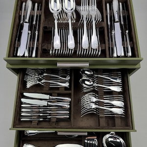 Può includere: Un set di posate argentate, tra cui cucchiai, forchette e coltelli, ordinatamente disposte in una scatola di legno verde. Le posate presentano un design decorativo sui manici. La scatola è aperta, rivelando più cassetti.