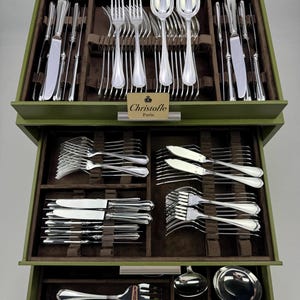Può includere: Un set completo di posate Christofle Paris in argento, ordinatamente disposto in una scatola di legno verde. Il set comprende forchette, coltelli, cucchiai e posate da portata. Il logo Christofle Paris è visibile su una piccola carta. Le posate sono in metallo argentato lucido.