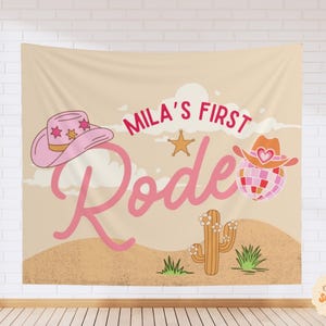 Puede incluir: Un tapiz de pared rectangular de tela con un paisaje des&eacute;rtico beige, un cactus y el texto Mila's First Rodeo en letra cursiva rosa. El dise&ntilde;o incluye un sombrero de vaquero rosa con estrellas, una estrella dorada y una bola de discoteca.