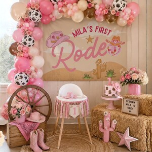 Puede incluir: Un conjunto de decoraci&oacute;n de fiesta con una pancarta con el texto Mila's First Rodeo, un arco de globos rosa y blanco, una trona con una pancarta de n&uacute;mero uno, un pastel con estampado de vaca, una rueda de madera, botas vaqueras y heno.