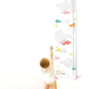 May include: A white growth chart with a colorful cloud and star design. The chart is hanging on a white wall and a child is standing next to it. The chart has a measurement scale in inches and the name "Olivia" is printed at the bottom.