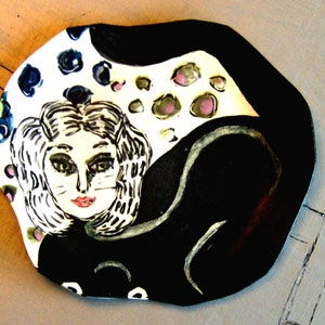 Può includere: Un piatto in ceramica con un design stravagante. Il piatto presenta il volto di una donna con tratti felini, in bianco e nero. Una coda di gatto nera si curva attorno alla donna. Lo sfondo presenta macchie colorate.
