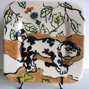 Puede incluir: Un plato cuadrado de cerámica blanca con un diseño pintado a mano de un perro blanco y negro con follaje marrón y verde en el fondo. El plato está sobre un soporte de metal negro.