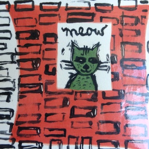 May include: A square ceramic tile with a hand-painted design. The central image features a green cat with the word "meow" above it. The background has a red and black geometric pattern.