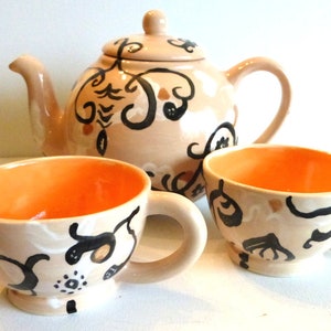 May include: A beige teapot and two teacups with black and orange floral designs. The teapot has a handle and a spout. The teacups have handles and are filled with orange liquid.