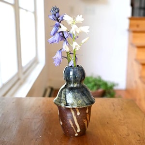 May include: A small ceramic bud vase with a bulbous neck and a tapered base. The upper section features a dark blue and black glaze, while the lower body is a textured brown with vertical white drip patterns, holding blue and white bell-shaped flowers.