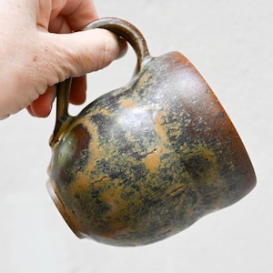 May include: A handmade ceramic mug with a unique, textured surface. The mug features a dark brown and olive green mottled glaze, with a sturdy handle. The mug is held by a hand, showcasing its size and shape. Perfect for coffee or tea.